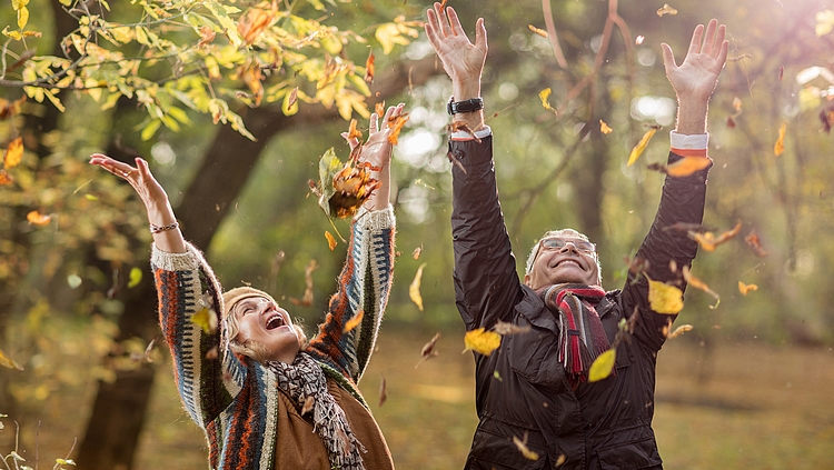 Två seniorer, en kvinna och en man, står utomhus. Det är en solig höstdag och de kastar löv upp i luften och skrattar.
